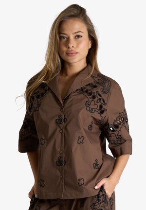 Donna con capelli lunghi che indossa una camicia marrone con bottoni e pantaloni abbinati con motivi ricamati neri ritagliati.