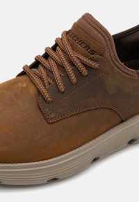 Braune Ledersneaker mit strukturierten Oberflächen, braunen Schnürsenkeln und einer gepolsterten grauen Sohle. Verfügt über ein Logo auf der Zunge und gestickte Akzente.