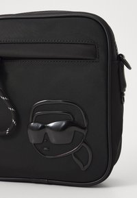 Black nylon shoulder bag featuring a glossy black abstract face design with sunglasses and a textured strap. Zippered pocket on the back.