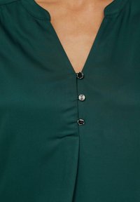 Ciemnozielona bluzka z dekoltem w kształcie litery V, pięcioma metalowymi guzikami o teksturowanym wzorze oraz gładkim, lekkim materiałem.