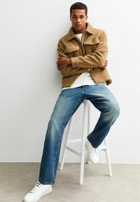 Bruine fleecejas met een kraag en twee voorzakken, gecombineerd met blauwe rechte jeans en witte sneakers. Minimalistisch ontwerp, neutrale kleuren.