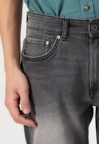 Jeans en denim gris avec une texture délavée, cinq poches et une fermeture à bouton au niveau de la taille, tenus par une main près de la hanche.