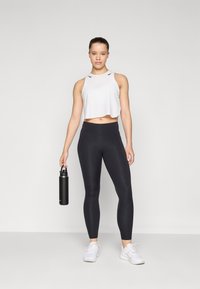 Débardeur blanc court avec des accents noirs, legging noir taille haute, tenant une bouteille d'eau noire, portant des chaussures de sport blanches.