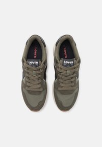 Baskets vert olive avec une combinaison de matériaux en tissu et en daim, avec des accents noirs et une semelle texturée. Nom de la marque visible sur la languette.