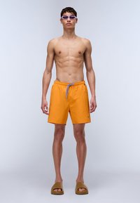 Shorts de bain orange vif avec ceinture élastique et cordon de serrage bleu, associés à des sandales marron et des lunettes de soleil teintées de violet.