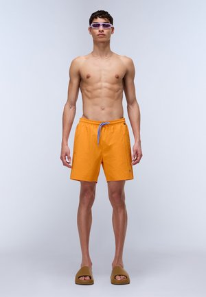 Bañadores de color naranja brillante con cintura elástica y cordón azul, combinados con sandalias marrones y gafas de sol con tinte púrpura.