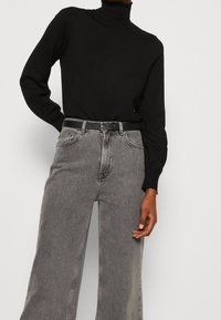 Schwarzer Rollkragenpullover kombiniert mit hoch taillierten, weiten grauen Jeans. Das Outfit verfügt über einen schwarzen Gürtel und dezente Stichdetails.