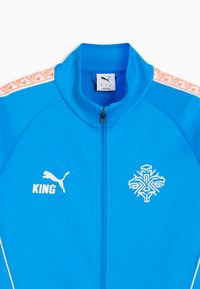 Veste de survêtement Puma bleu vif avec texte "KING" blanc et logo sur le côté gauche de la poitrine, emblème géométrique blanc sur le côté droit de la poitrine, et bandes d'épaule orange à motif.