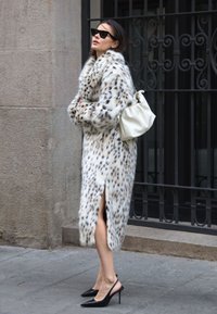 Hosszú fehér műszőrme kabát fekete foltokkal, oldalsó hasítékkal, fekete magassarkú szandálokkal viselve. Fehér kézitáskával párosítva. Háttérként egy textúrált fal szolgál.