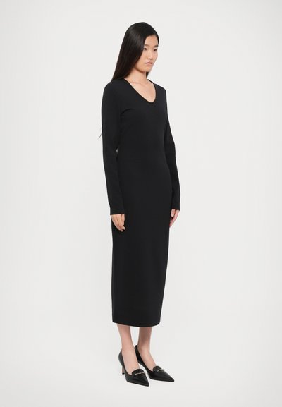 Black long-sleeve dress with a V-neck, made of a smooth fabric. The dress has a straight silhouette, reaching mid-calf, paired with pointed black shoes.