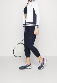 Biała i granatowa kurtka zapinana na zamek z paskami na mankietach, granatowe legginsy oraz szare buty do tenisa z czerwonymi akcentami. Trzyma rakietę tenisową.