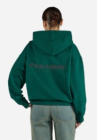 Moletão oversized verde feito de algodão, apresentando um grande logo nas costas em cinza escuro. Inclui capuz com cordão e punhos canelados.