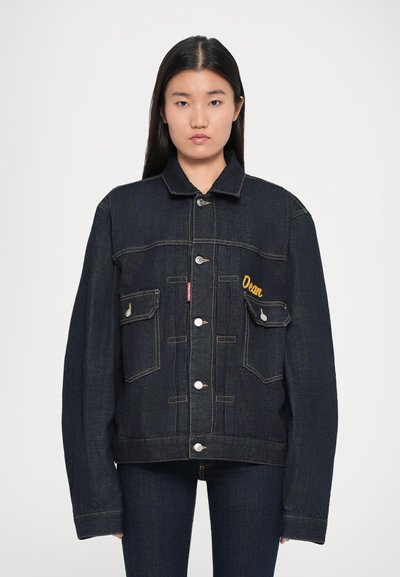 Mørk denimjakke med knaplukning, to brystlommer og gul broderi. Har vide ærmer og kontrasterende syninger.