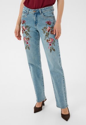 Femme portant un jean bleu clair avec une broderie florale rose sur les cuisses, un haut bordeaux et des chaussures à talons marron, debout.