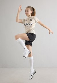 Sportig outfit med en beige randig tröja med "SAVVY"-märke, svarta shorts, vita knästrumpor och vita fotbollsskor med svarta detaljer.