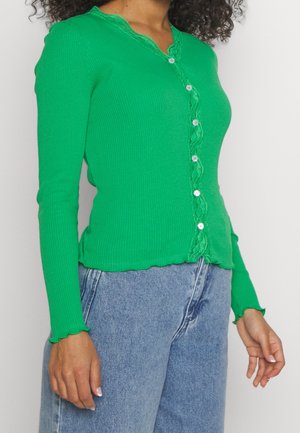 Frau trägt eine leuchtend grüne, gerippte Strickjacke mit Knöpfen und Spitzenbesatz, kombiniert mit hoch taillierten hellblauen Jeans.