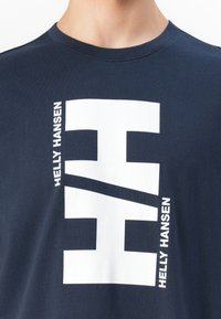 Dunkelblauses Hemd mit großem weißen Helly Hansen Logo und Markennamen, der vertikal auf der Brust aufgedruckt ist.