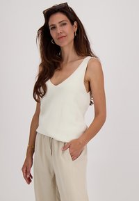 Witte mouwloze top met een V-hals, gemaakt van gestructeerde stof. Gecombineerd met beige, trekkoord broek. Model staat met één hand in de zak.