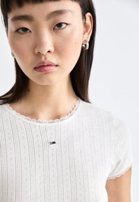 Hvit ribbestrikket topp med korte ermer, blondekant ved halslinningen, med en liten logo i rød, hvit og blå nær midten.