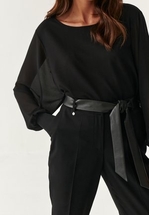 Kobieta ubrana w czarną bluzkę z długim rękawem z przezroczystymi rękawami oraz czarne spodnie z wysokim stanem, związane szerokim skórzanym paskiem.