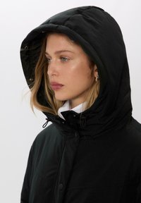 Čierna vypchatá bunda s vysokým golierom a priliehavou kapucňou. Obsahuje stiahnutia, cvoky a hladkú textúru.