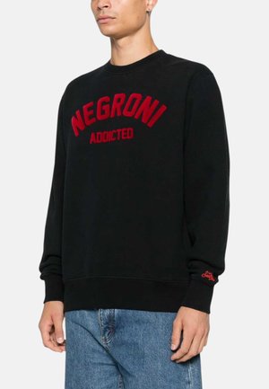 Uomo che indossa una felpa nera con scritta rossa "NEGRONI ADDICTED" e jeans blu, in piedi contro uno sfondo chiaro e uniforme.