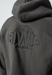 Tamnosiva majica s kapuljačom s teksturiranim izvezenim logotipom s natpisom "RIVWIA PROJECTS" na leđima. Mekani materijal s dizajnom kapuljače.