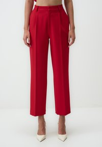 Red tailored trousers with a high waist, pleated front, straight legs, and a smooth fabric texture. Worn with white pointed heels.