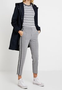 Parka à capuche marine sur un haut rayé blanc et marine, associée à un pantalon à carreaux avec des rayures latérales et des baskets blanches.