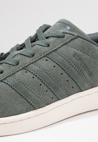 Zapatilla de ante verde con seis franjas, superficie texturizada y perforaciones. Presenta una suela de goma blanca y un acento brillante de marca.