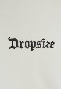 Tissu blanc présentant le mot "Dropsize" en broderie gothique noire en gras, mettant en avant le contraste entre le tissu et le texte.