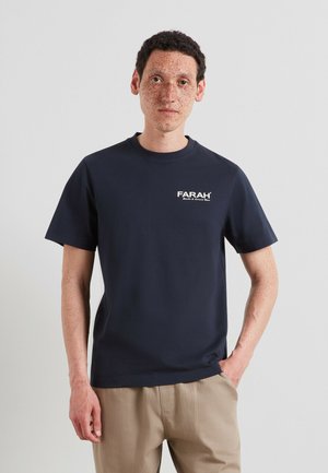 Uomo con capelli corti e ricci e lentiggini, che indossa una maglietta Farah blu navy e pantaloni beige, in piedi davanti a uno sfondo chiaro uniforme.