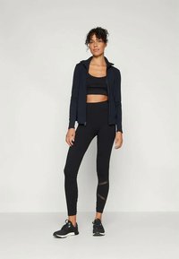 Ensemble de sport noir composé d'une veste zippée, d'un top court et de leggings taille haute avec des accents en mesh, complété par des baskets noires.