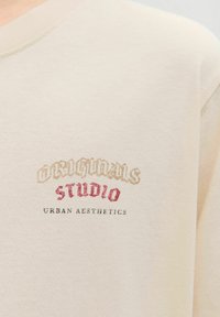 Pūkais džemperis ar apaļu kakla izgriezumu, ar izšūtu tekstu "ORIGINALS STUDIO" zeltā un sarkanā krāsā, un "URBAN AESTHETICS" zemāk pelēkā krāsā.