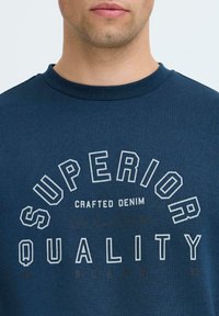Sudadera de cuello redondo en azul marino, fabricada en un material suave, con un diseño impreso que dice "CALIDAD SUPERIOR" en letras blancas y beiges.
