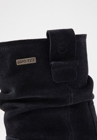 Botín de ante negro con un diseño plegable, presenta una etiqueta de metal con la inscripción "DUO-TEX" y un detalle de costura en el lateral.