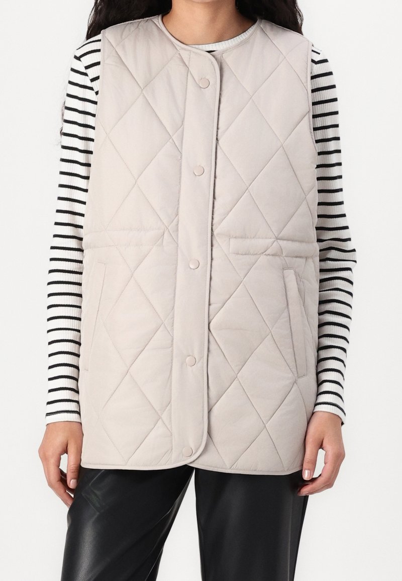 Femme portant un gilet matelassé beige sans manches avec des boutons-pression, par-dessus une chemise à manches longues rayée noir et blanc et un pantalon noir.