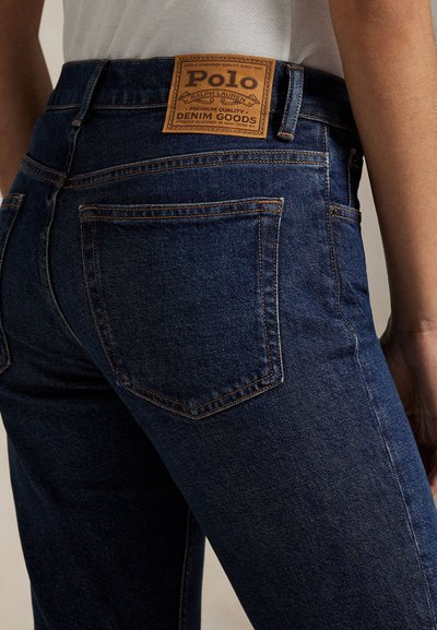 Ingrandimento di una persona che indossa jeans in denim blu scuro con il distintivo in pelle del marchio Polo Ralph Lauren sopra la tasca posteriore.