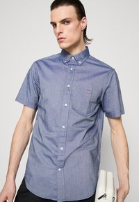 Młody mężczyzna z zaczesanymi do tyłu włosami, ubrany w krótki niebieski koszulowy shirt, trzymający w lewej ręce białą saszetkę na zamek.