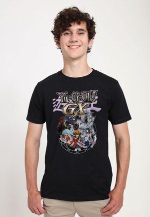 Jonge man die lacht, draagt een zwart T-shirt met kleurrijke Yu-Gi-Oh! GX animefiguren en logo op de voorkant, beige broek, staande tegen een witte achtergrond.