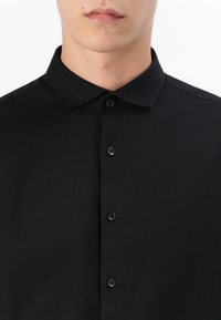 Primer plano de una persona que lleva una camisa negra abotonada con cuello puntiagudo sobre un fondo blanco liso.