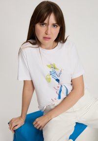 Biała bawełniana koszulka z nadrukiem przedstawiającym postać w niebiesko-zielone paski, z napisem "le plage" w kolorze czerwonym, noszona z jasnobeżowymi spodniami.