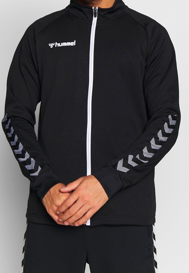 Homme portant une veste de sport noire zippée avec une fermeture éclair blanche, logo Hummel sur la poitrine, et motifs chevrons réfléchissants sur les manches.