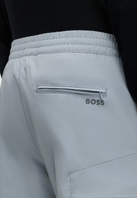 Pantalon gris clair avec taille élastique, poche arrière zippée et logo BOSS près de la poche sur une personne portant un haut noir.