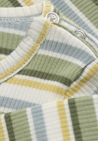 Geribbelde, gemêleerde stof met strepen in groen, geel en blauw. Bevat drukknoopsluitingen nabij de kraag voor eenvoudig aan- en uittrekken.
