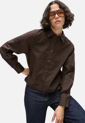 Personne aux cheveux bouclés et foncés portant une chemise marron oversize avec des patches texturés et un jean bleu foncé, posant avec la main sur la hanche et des lunettes teintées orange.