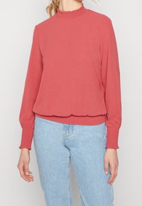 Koralli pitkähihainen paita, jossa on röyhelöinen korkeakaulus ja kerätty hihansuu. Pehmeä, joustava kangas, jossa on väljä istuvuus, yhdistettynä vaaleansinisiin farkkuihin.