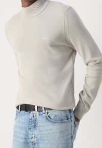 Pull col roulé beige clair avec col et poignets côtelés, comprenant des coutures latérales et un détail subtil du logo. Associé à un jean en denim bleu.