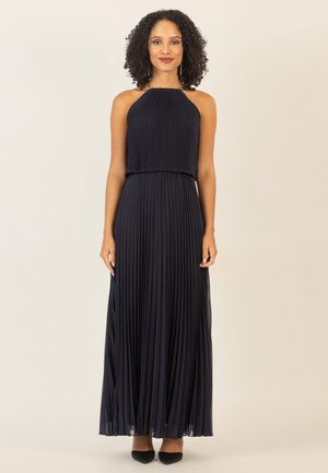 Robe maxi plissée marine avec décolleté halter, tissu texturé, silhouette fluide et détails subtils en transparence au niveau de l'ourlet. Talons aiguilles noirs.