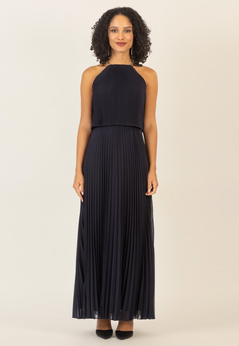 Vestido maxi de navy con plisados, escote halter, tejido texturizado, silueta fluida y sutil detalle traslúcido en el dobladillo. Tacones negros de punta.